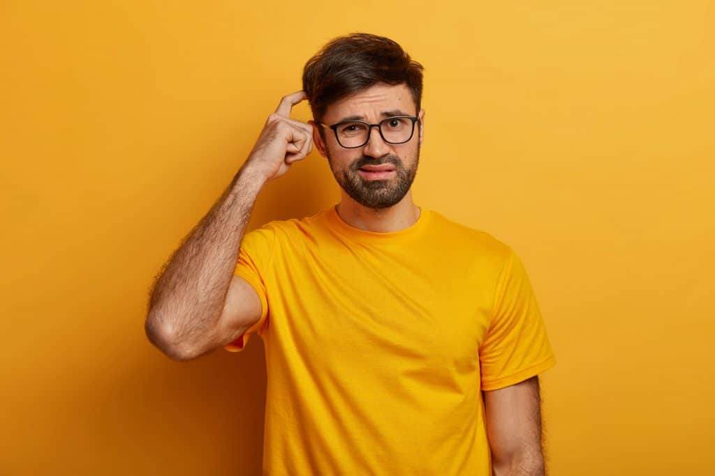 A man wearing a yellow shirt and glasses scratching his head in confusion.