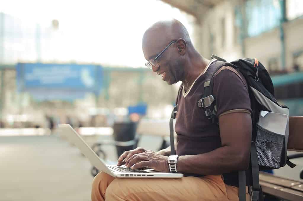 A smiling man using a VPN for expats, sat on a bench in a train station typing on his laptop.