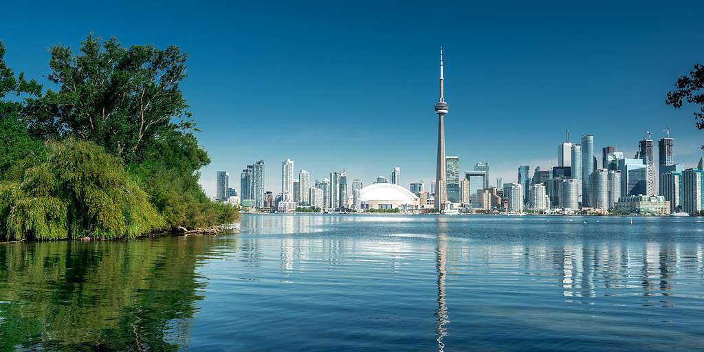 An image of Toronto, Canada. The city can be seen in the distance, mirrored by the reflection in the water. The CN Tower is the focus on the image. On the left, in the foreground, is a small, green island overflowing with bushes and trees.