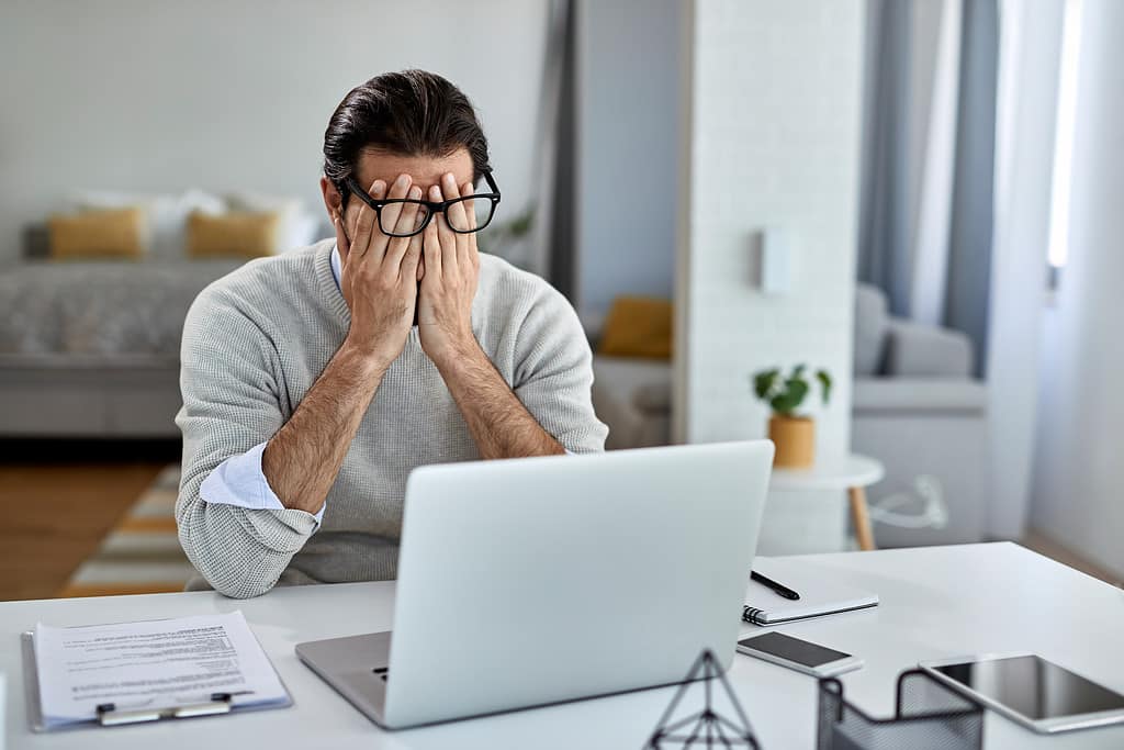 A man, sitting at a laptop, sitting rubbing his eyes with frustration. He has been caught by a legalisation scam.