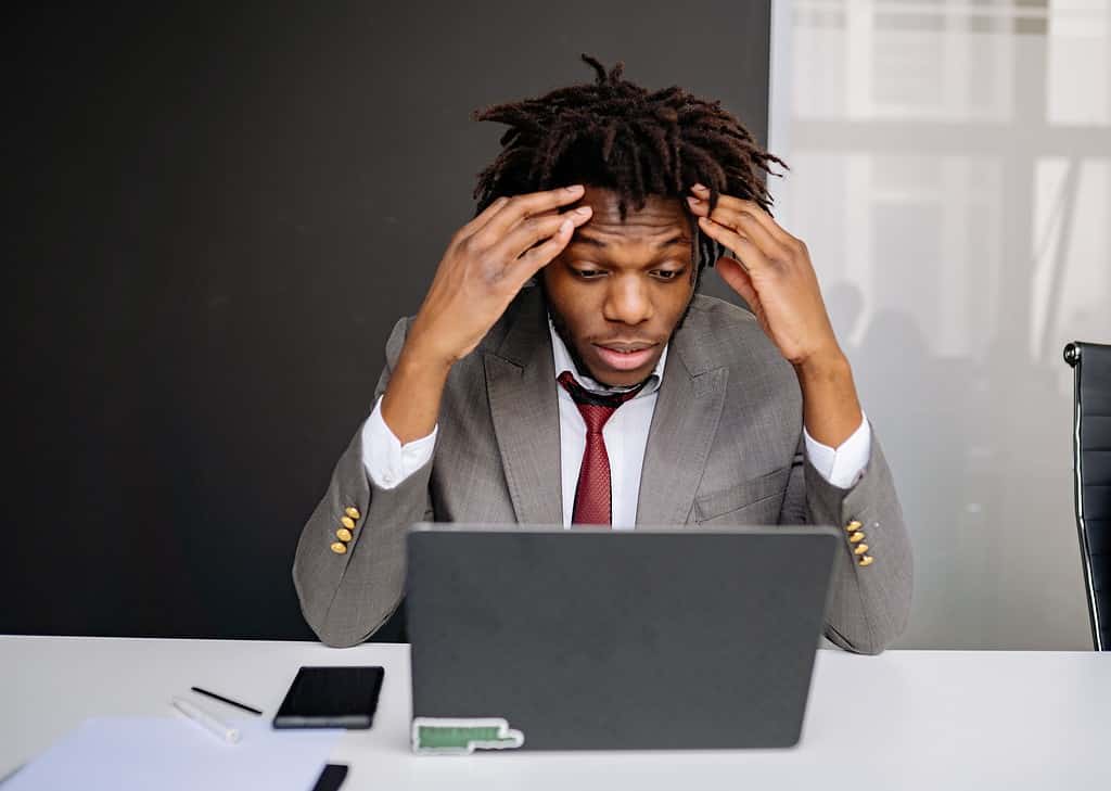 A businessman in a suit sat in front of his laptop with his head in his hands.