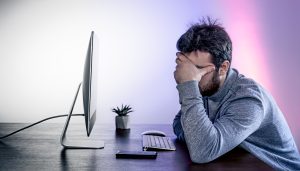 tired man sits front computer covering his face with his hands