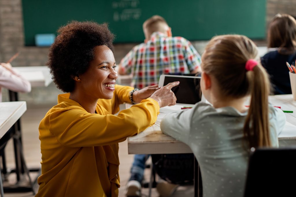 A smiling teacher pointing to an iPad, knelt down at one of her student's desks.