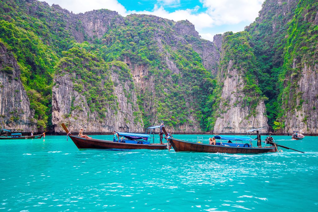 An image of a lagoon with striking cliffs in Thailand, covered in green shrubbery. In the foreground two boats in vibrant blue water, for Thailand joining the Hague blog.